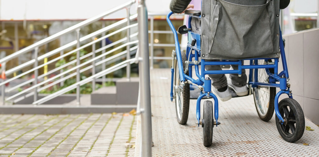 A man in a blue wheelchair is moving up an accessible wheelchair ramp toward a building entrance.